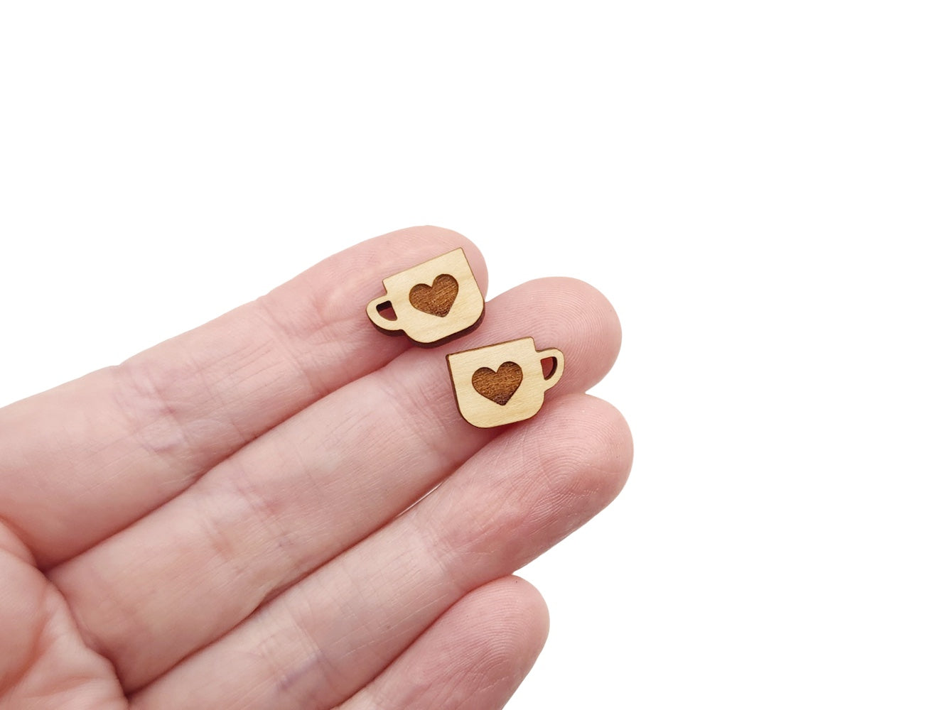 a hand holding a pair of wooden cabochon stud earring blanks cut and engraved to look like a mug with a heart