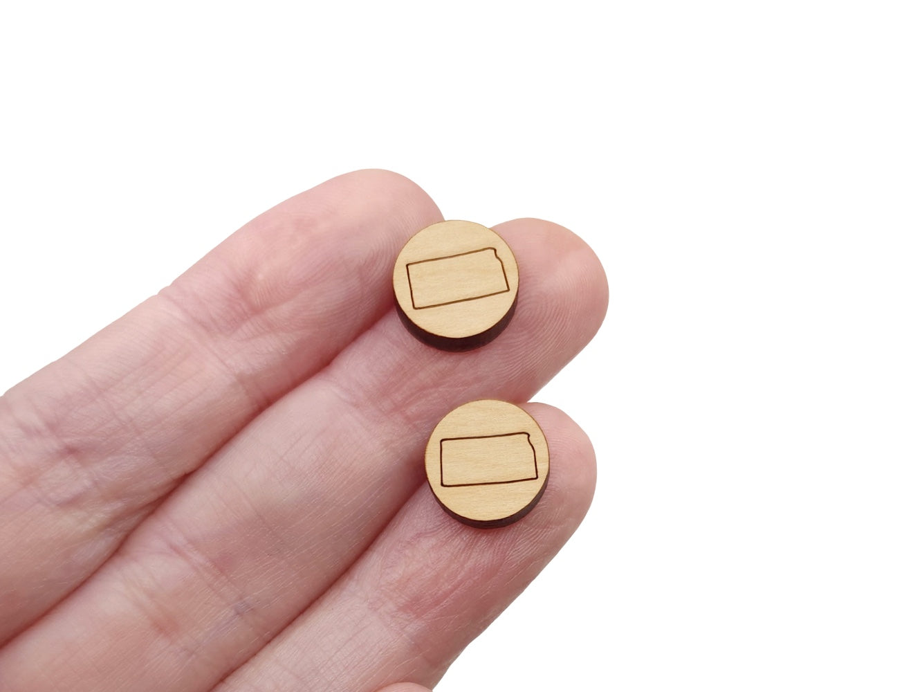 a hand holding a pair of round wooden cabochon earring blanks engraved with the state of Kansas outline