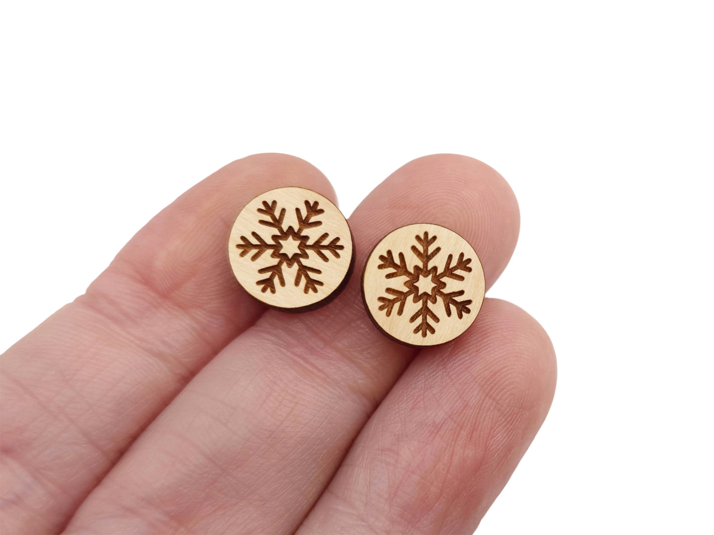 a hand holding a pair of round wooden cabochon earring blanks engraved with a snowflake