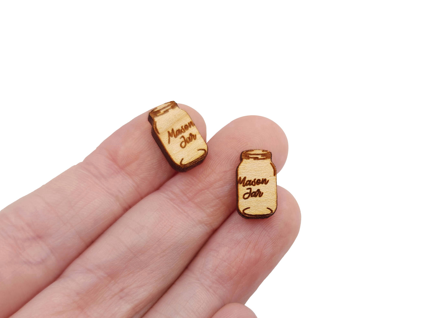 a hand holding a pair of small wooden cabochon blanks for stud earrings, cut and engraved to look like mason jars