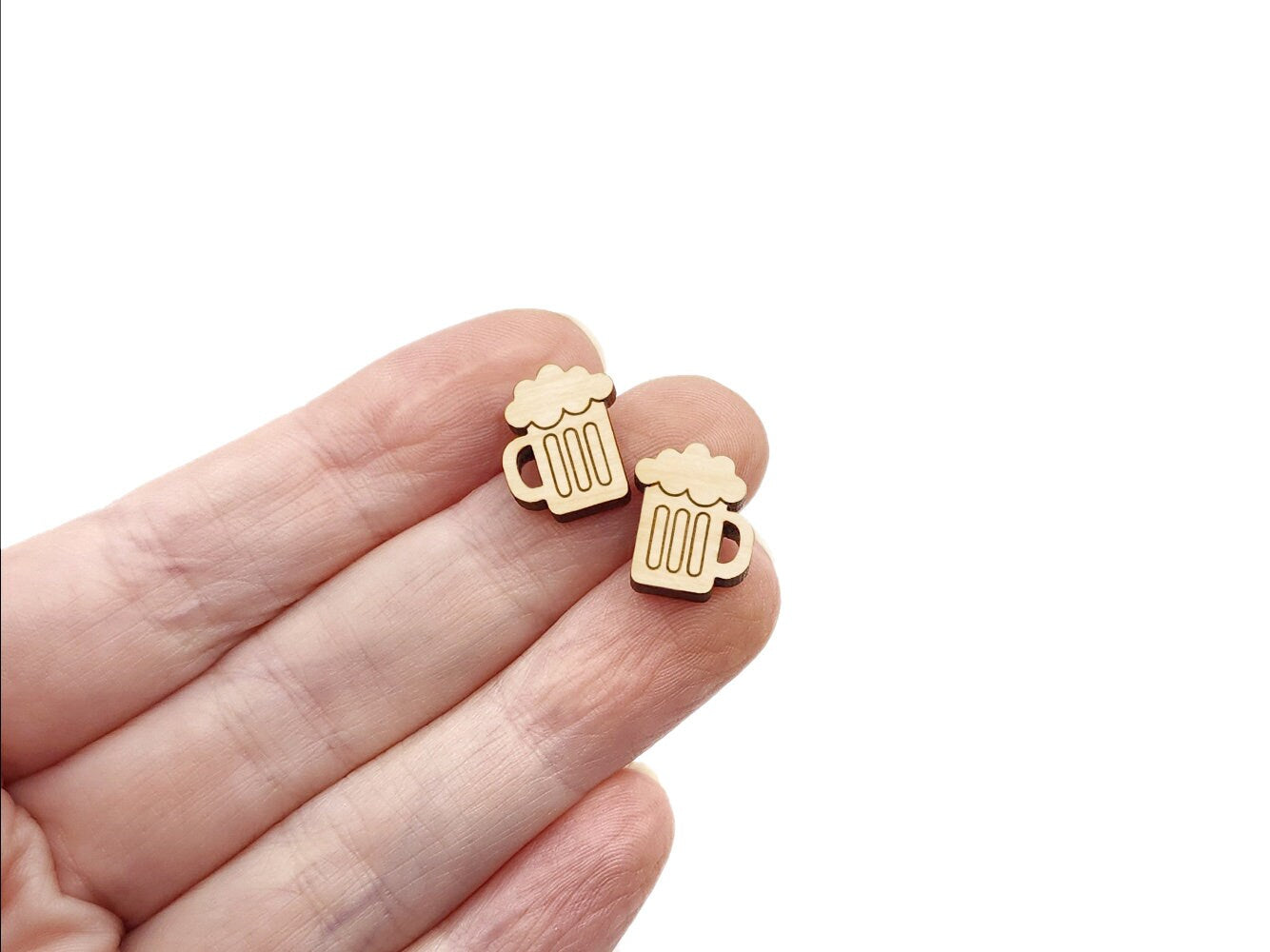 a hand holding a pair of wooden cabochon stud earring blanks cut and engraved to look like mugs of beer