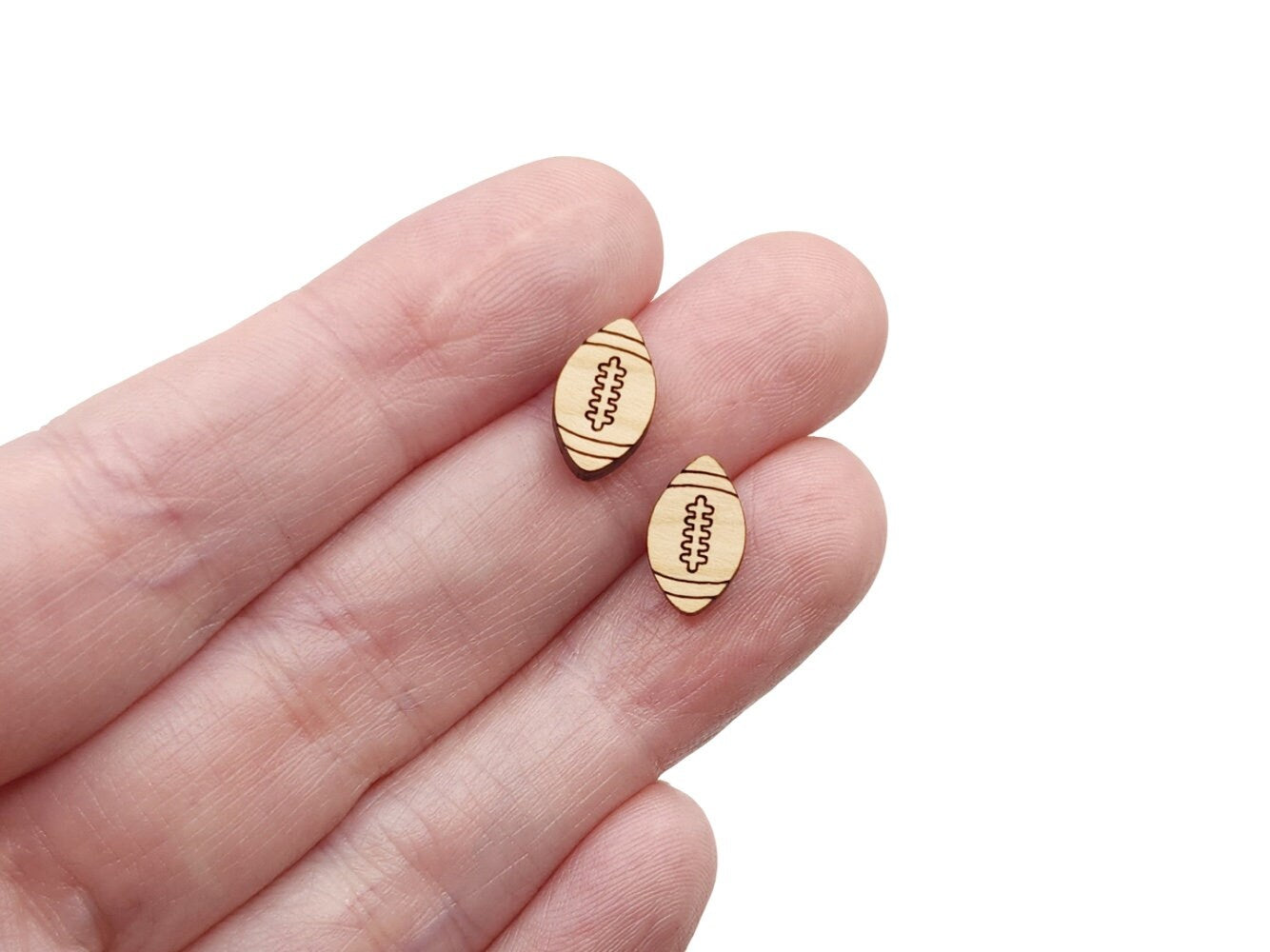a hand holding a pair of wooden cabochon stud earring blanks cut and engraved to look like footballs