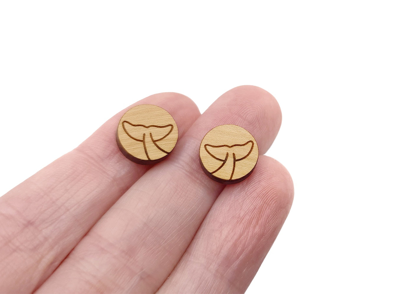 a hand holding a pair of round wooden cabochon earring blanks engraved with a whale's tail