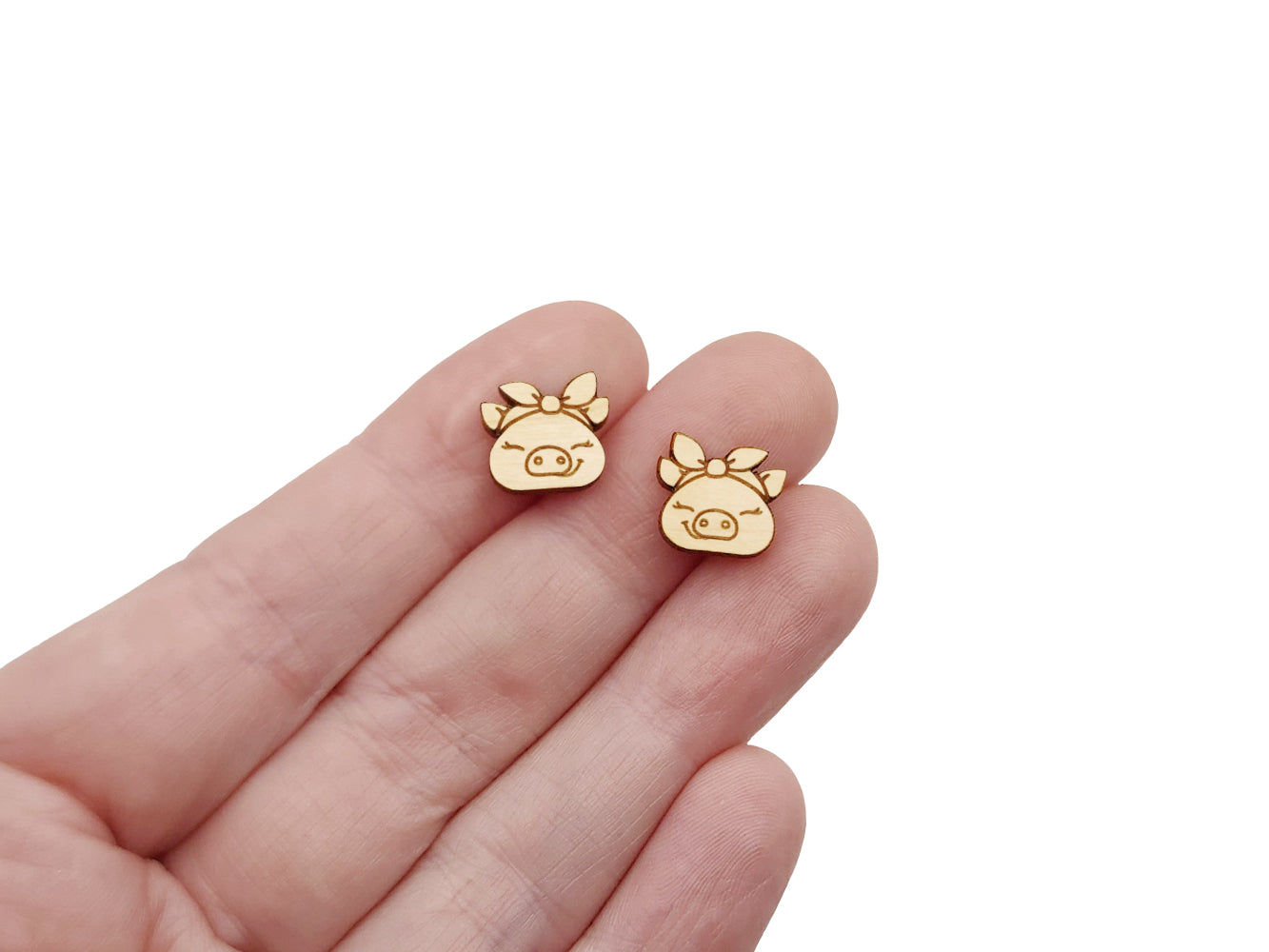 a hand holding a pair of wooden cabochon stud earring blanks cut and engraved to look like pigs wearing a bandana