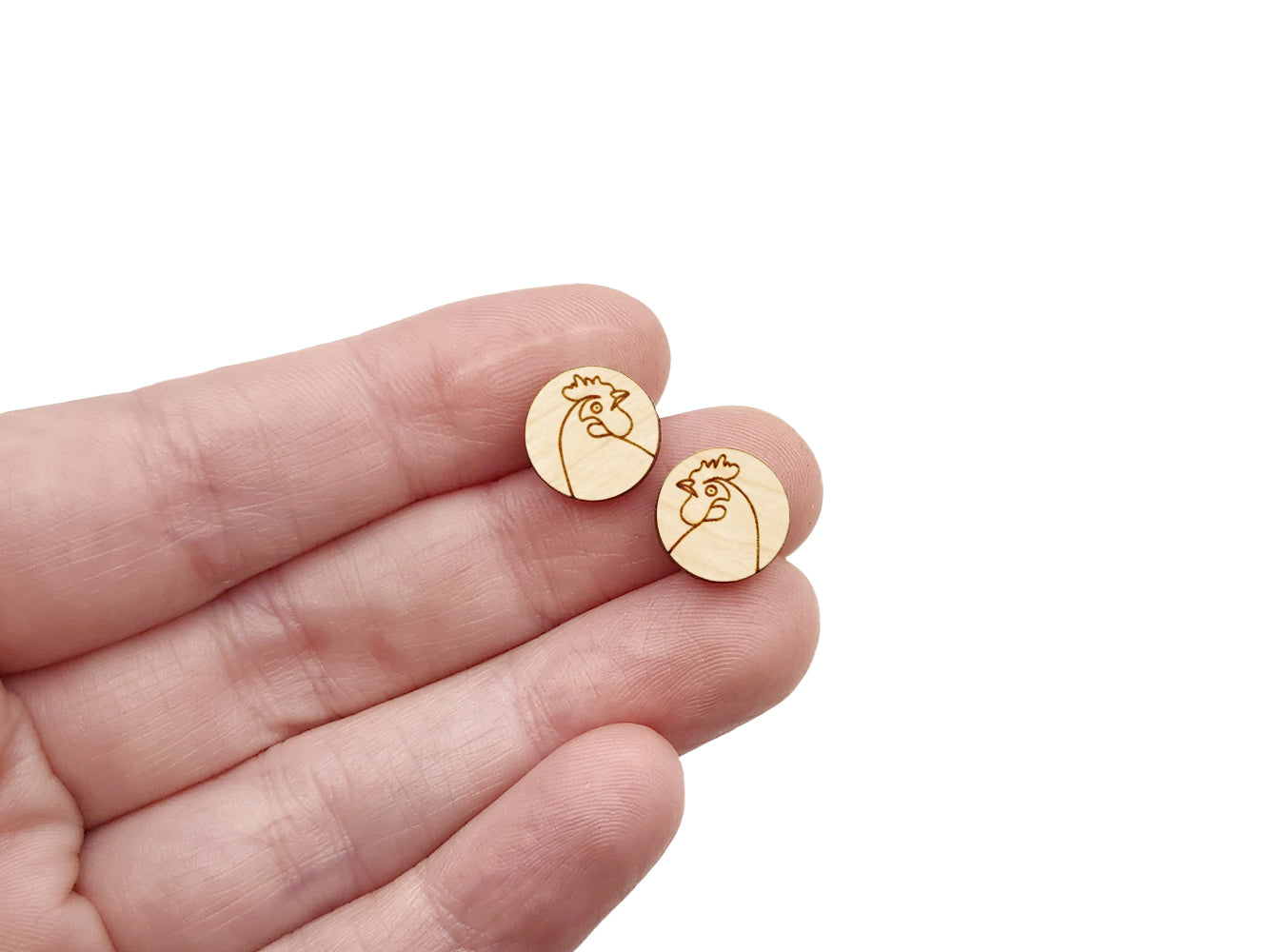 a hand holding a pair of round wooden cabochon earring blanks engraved with a chicken