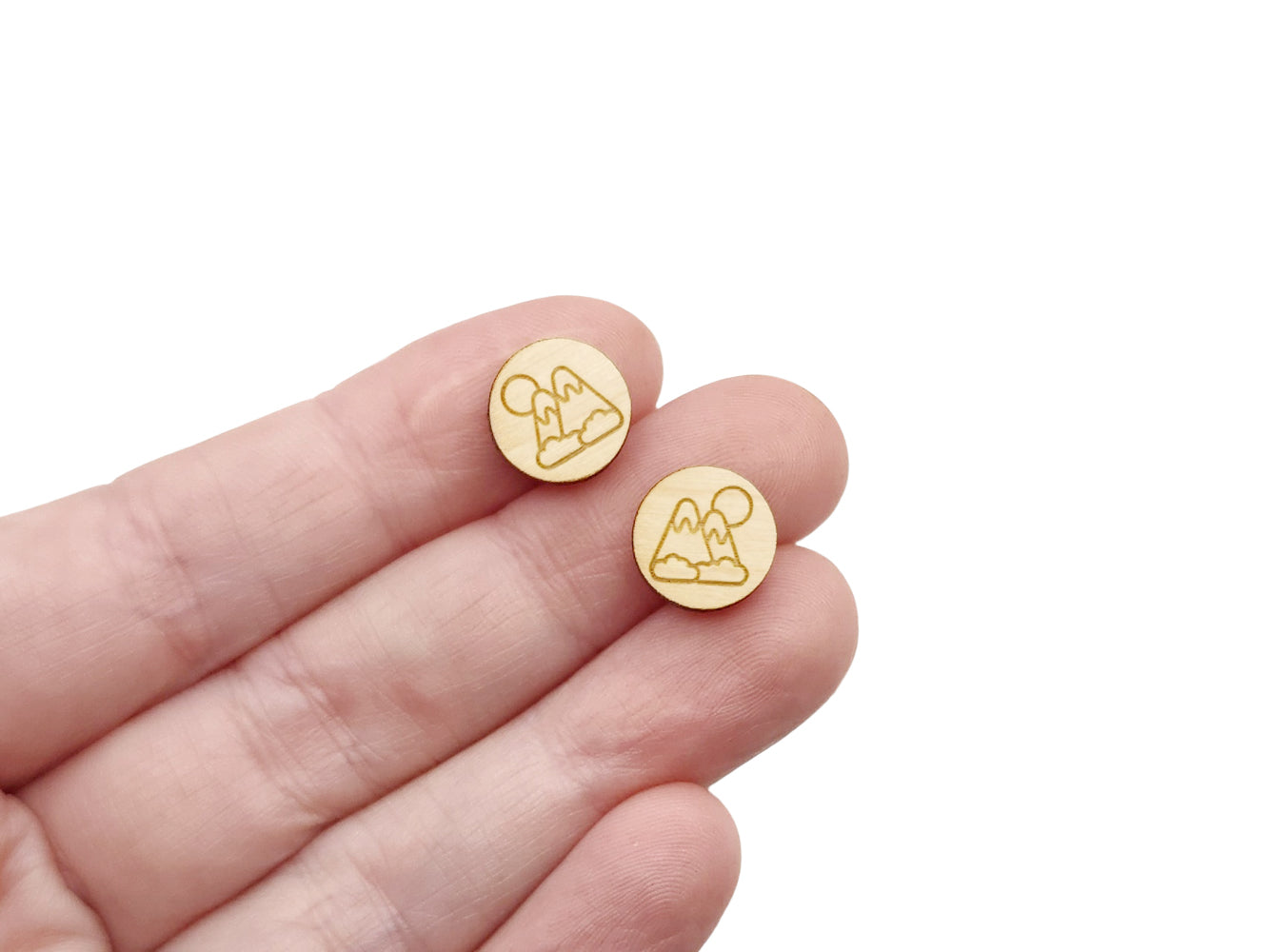 a hand holding a pair of round wooden cabochon earring blanks engraved with a mountain scene
