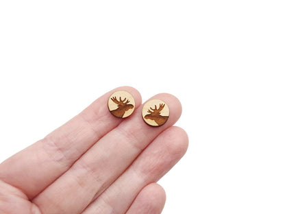 a hand holding a pair of round wooden cabochon stud earring blanks engraved with a moose silhouette