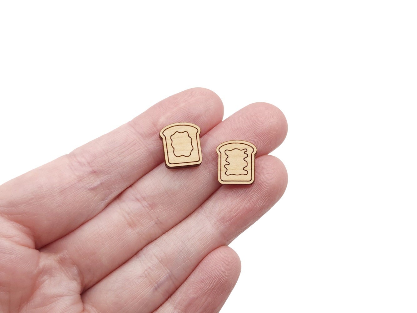 a hand holding a pair of wooden cabochon stud earring blanks cut and engraved to look like pieces of bread with peanut butter and jelly